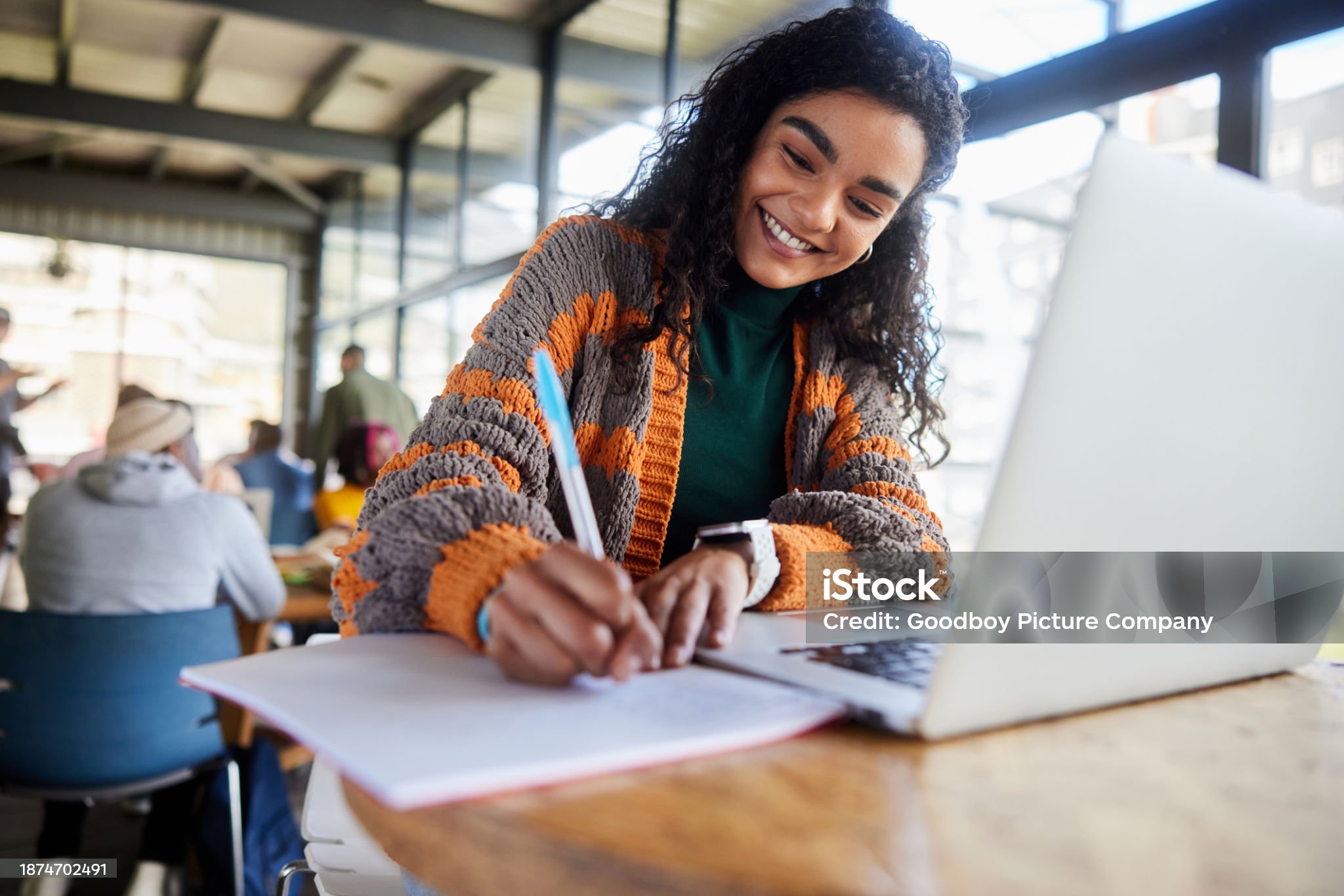 Low angle view of a smiling young female college student writing notes and working on a laptop while doing her homework in a busy campus cafeteria
