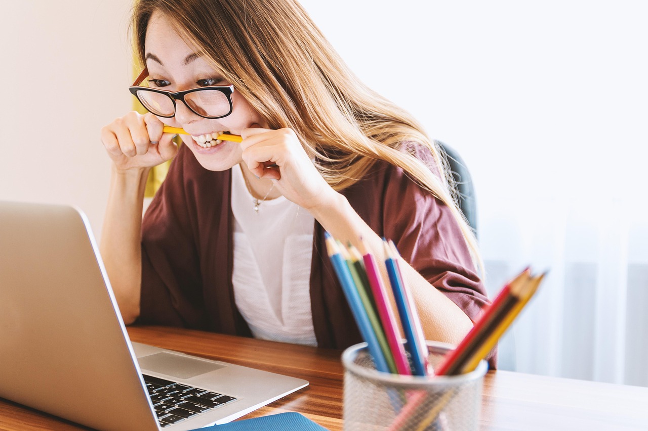 lady looking at laptop and biting down a pencil, there is a small container of colored pencils on the table